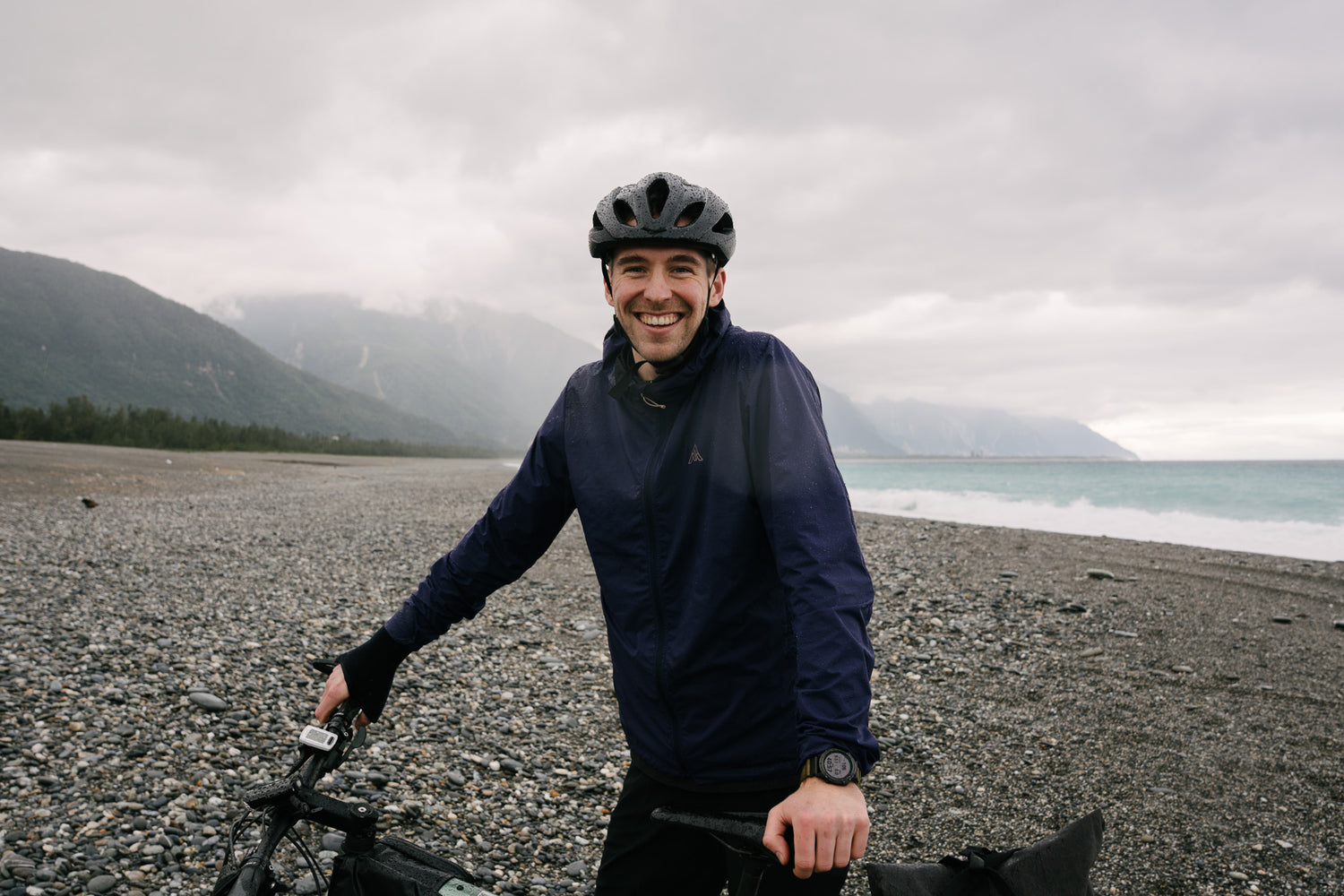 AYR Cycling founder smiling during a bikepacking trip on a rocky coastal beach under cloudy skies